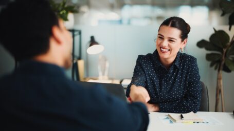 Photo of a smiling woman behind a desk shaking hands with someone in an office setting.